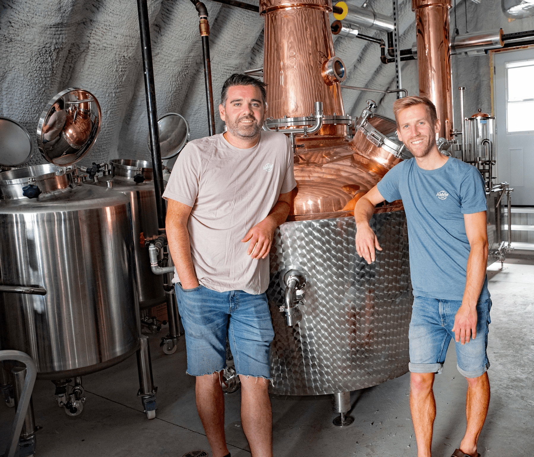 Ryan & Taylor standing in a brewery with large copper and stainless steel tanks.