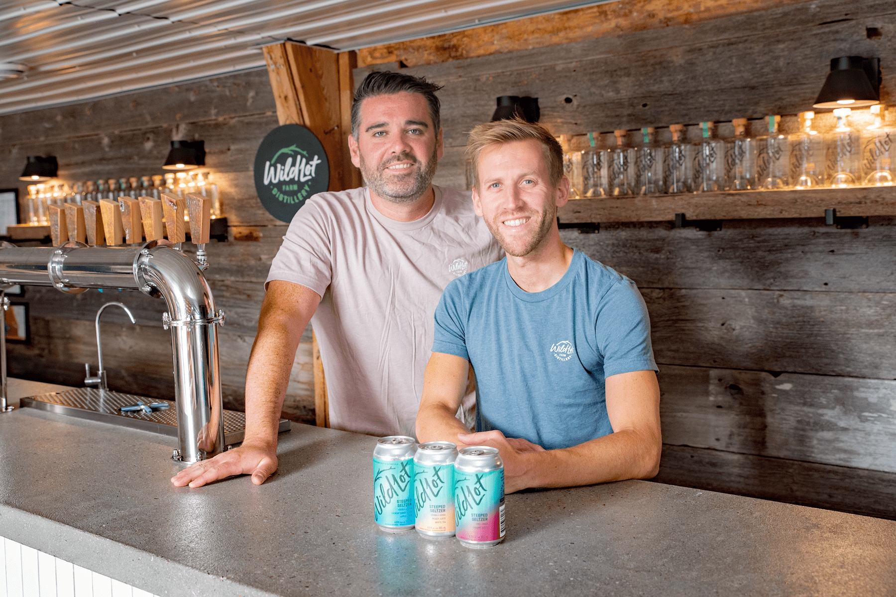 Ryan & Taylor standing behind a bar with cans of beer in front of them.