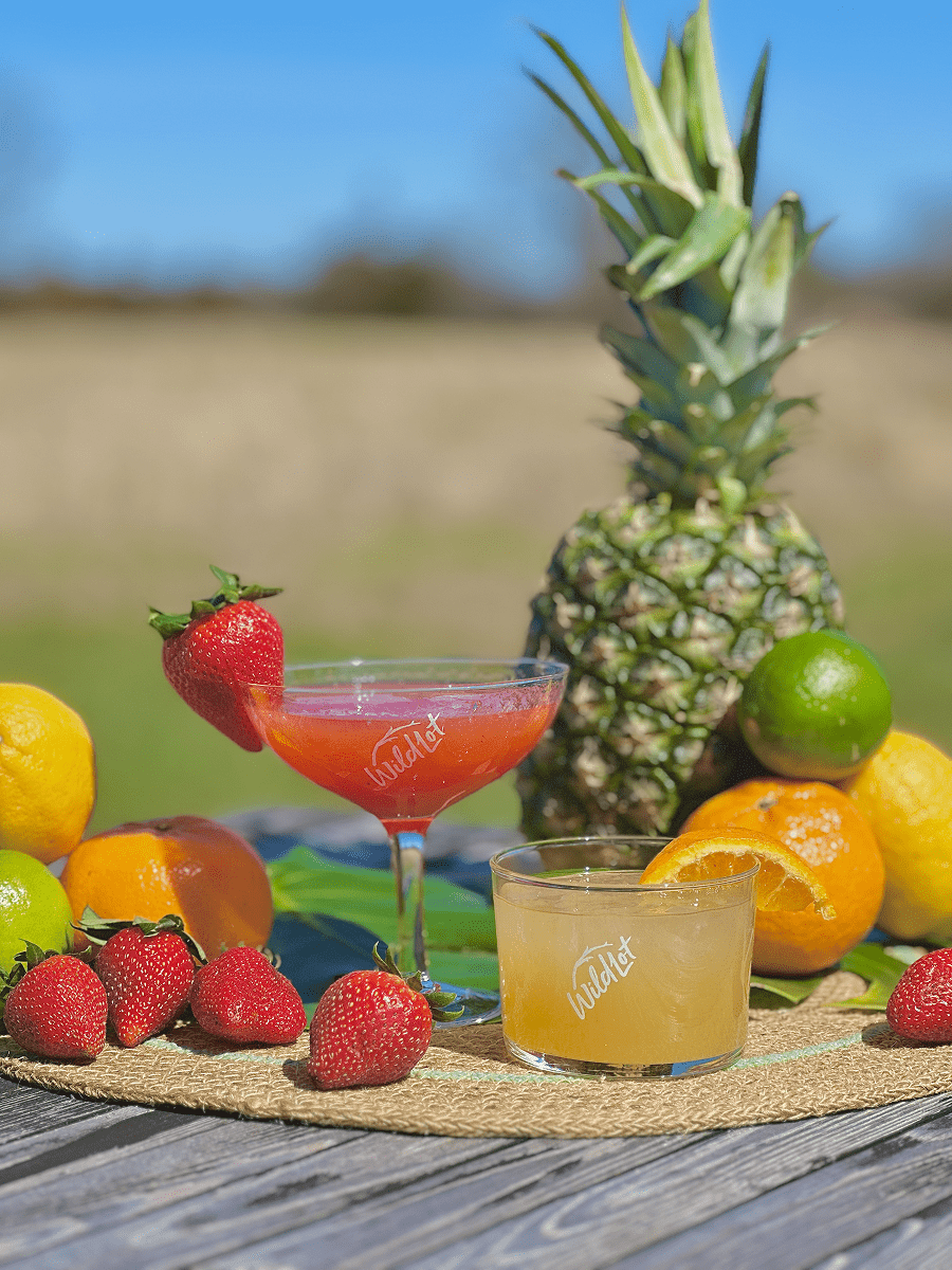 Assorted fruits including a pineapple, lemons, limes, and strawberries with two drinks on a wooden surface.