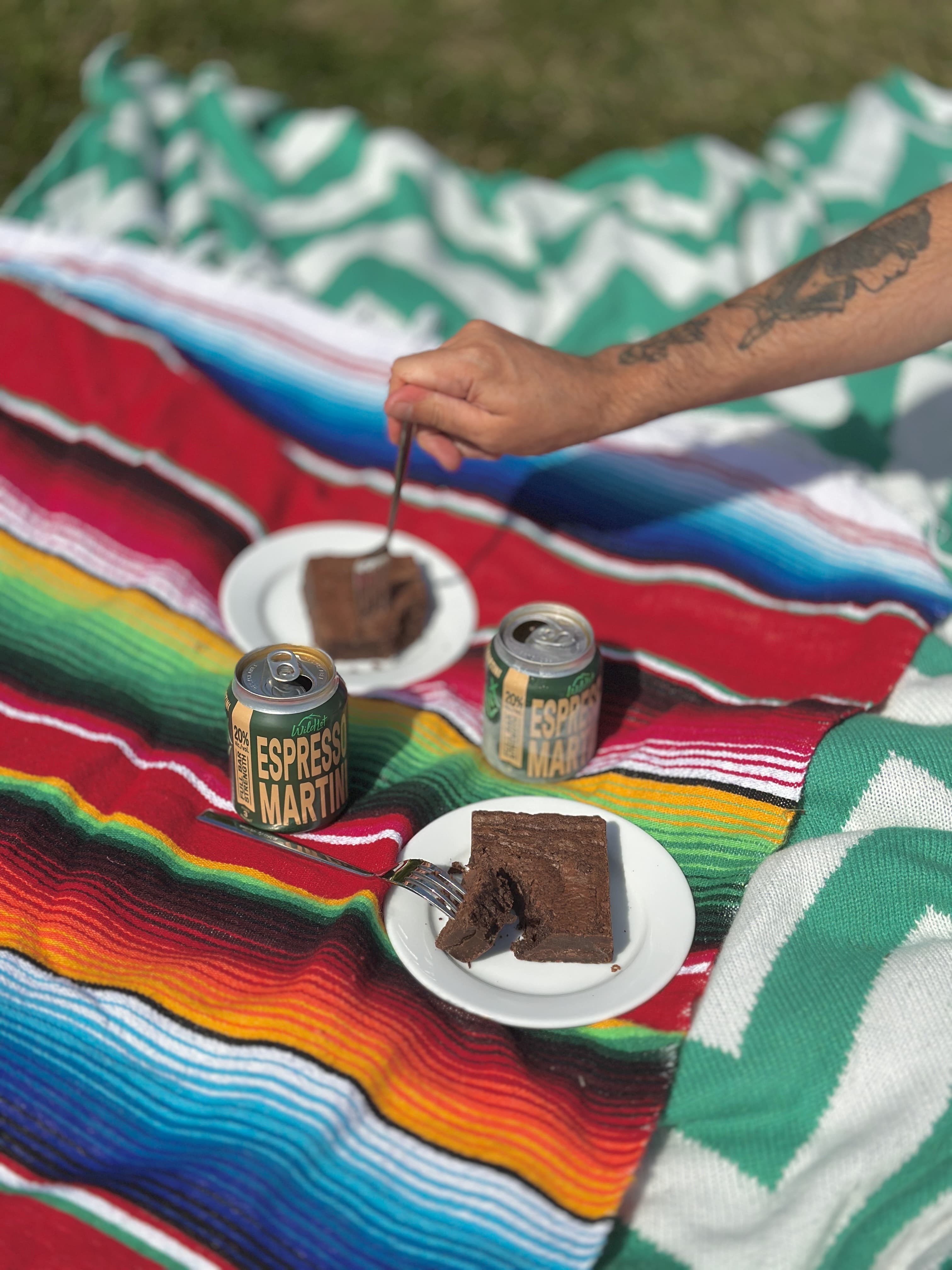 Person enjoying a slice of chocolate cake with espresso martini on a colorful blanket outdoors.